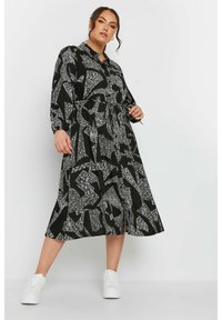 Abito midi nero con motivi astratti bianchi, maniche lunghe, chiusura a bottoni, cordoncino in vita e spacco laterale. Abbinato a sneakers bianche.