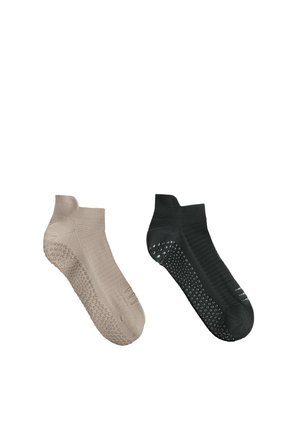 Une chaussette de cheville beige et une noire, chacune avec des grips en caoutchouc antidérapants sous la semelle, exposées sur un fond blanc.