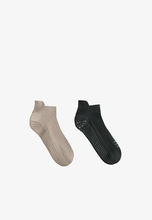 Une chaussette de cheville beige et une noire, chacune avec des grips en caoutchouc antidérapants sous la semelle, exposées sur un fond blanc.