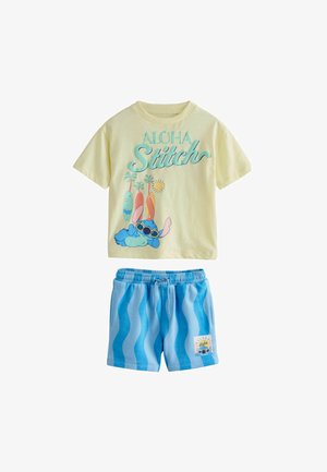 Žuta majica kratkih rukava s tekstom "Aloha Stitch", crtani Stitch s sunčanim naočalama i plave kratke hlače s valovitim prugama i malom zakrpom Stitch.