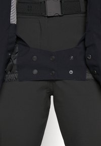 Pantaloni neri con texture liscia; presentano bottoni a pressione, una vita flessibile e una tasca nascosta in vita per una maggiore funzionalità.