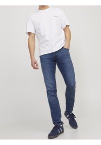 Muž oblečený v bílém tričku s krátkým rukávem, modrých slim-fit džínech a tmavě modrých teniskách s bílými pruhy, stojící proti jednoduchému šedému pozadí.