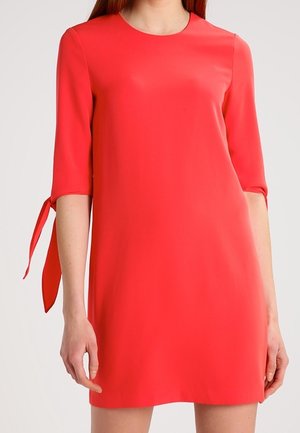 Kobieta ubrana w jaskrawoczerwoną sukienkę do kolan z rękawami do łokci i zawiązanymi kokardami na ramionach, stojąca na jednolitym tle.