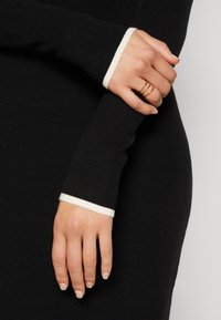 Robe en tricot noire avec des manches ornées d'un poignet blanc contrastant. Texture lisse avec des détails côtelés. Main ornée de fines bagues en or.