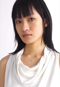 Guld-hänge på ett delikat kedja, med en cirkulär design. Modellen bär en vit topp med cowldesign i mjukt tyg och draperad textur.