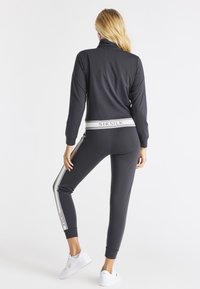 Survêtement noir comprenant une veste zippée avec un col haut et un pantalon assorti. Les deux ont une coupe slim et des rayures latérales contrastantes.