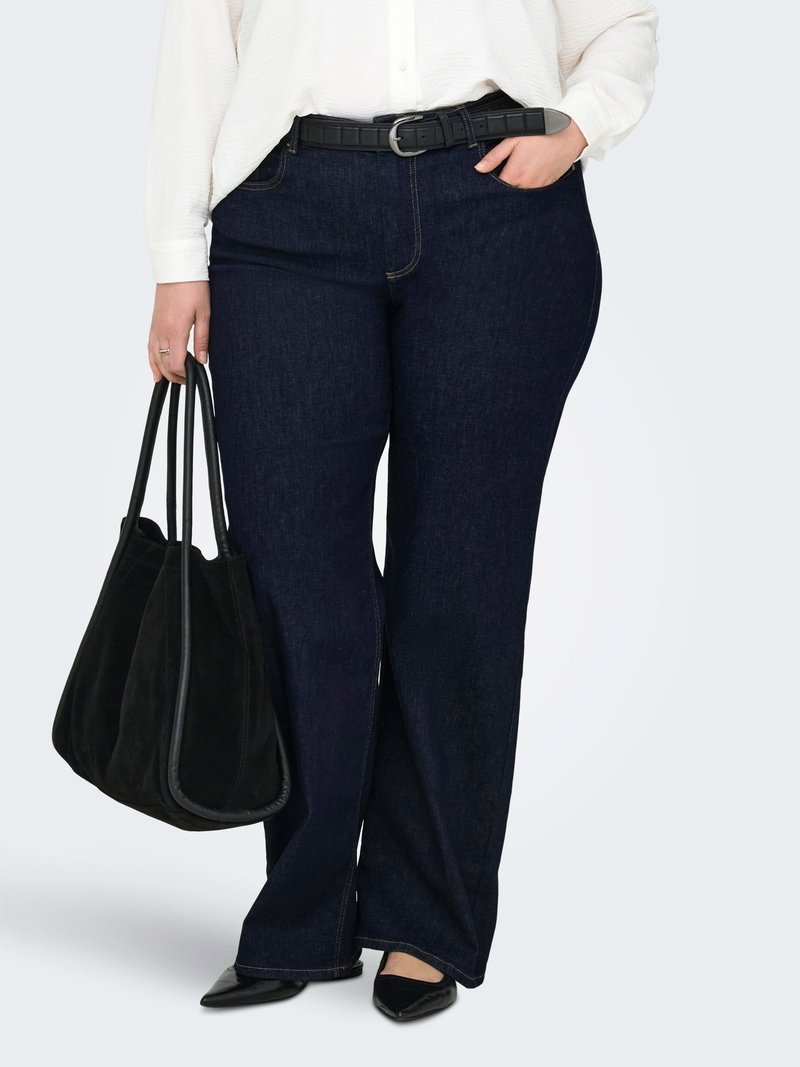 Femme portant un jean à jambes larges bleu foncé, une blouse blanche, une ceinture noire, des ballerines noires pointues, tenant un sac à main noir, une main dans la poche.