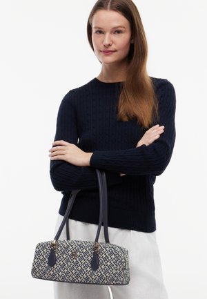 Vrouw met lang bruin haar die een navy kabelgebreide trui en witte broek draagt, een navy- en beige handtas met patroon en dubbele hengsels vasthoudend.