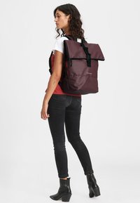 Sac à dos bordeaux avec un rabat, quincaillerie noire et bretelles réglables. Le matériau semble durable et résistant à l'eau, avec un design épuré.