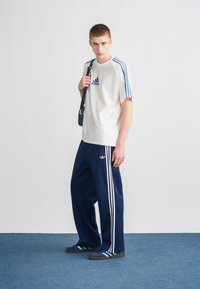 Adidas odjeća koja uključuje bijelu majicu s plavim akcentima i tamnoplave sportske hlače. Kompletnu odjeću čine plava obuća s tri pruge.