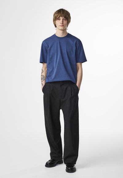 Mörkblå t-shirt med kort ärm och rund halsringning, matchad med svarta, veckade byxor. Modellen står med händerna i fickorna och har på sig svarta skor.