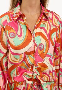 Blusa colorida con un vibrante patrón en espiral de color rosa, naranja y verde. Fabricada con una tela suave, cuenta con un cierre de botones en la parte delantera y un detalle de cintura atada.