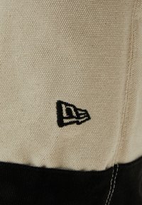 Tissu en coton crème avec une surface texturée, comportant une section de tissu noir et un logo noir brodé sur le bord.