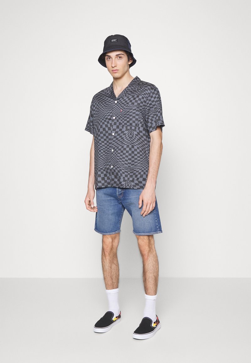 Hombre con un sombrero tipo cubo negro, camisa de cuadros gris de manga corta, pantalones cortos de mezclilla azules, calcetines blancos y zapatillas negras sin cordones con diseño de llamas.