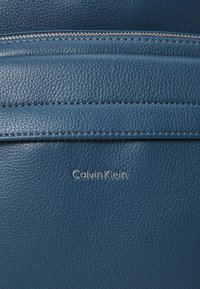 Material de cuero azul con un acabado texturizado. Presenta un zipper plateado y el logotipo "Calvin Klein" grabado en la esquina inferior derecha.