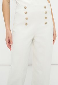 Γυναίκα που φοράει λευκό παντελόνι με υψηλή μέση και δύο κάθετες σειρές από τέσσερα χρυσά κουμπιά στην μπροστινή πλευρά, στέκεται μπροστά σε απλό φόντο.