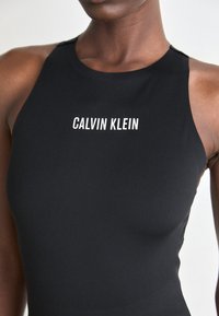 Zwarte mouwloze bodysuit met hoge halslijn, voorzien van witte "CALVIN KLEIN" tekst gecentreerd op de borst, gemaakt van gladde, elastische stof.