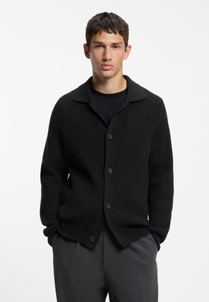 Ung mand iført en sort ribstrikket cardigan med knapper over en sort skjorte og mørkegrå bukser, stående mod en ensfarvet hvid baggrund.