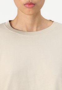 Beige katoenen t-shirt met een ronde hals, korte mouwen en een soepele textuur met subtiele stikdetails rond de kraag.