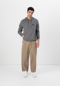 Hoodie cinza com bolso frontal, cordões e patch de logo. Combinado com calças beiges largas e ténis brancos.