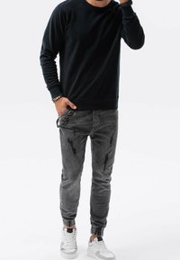 Felpa nera con collo rotondo abbinata a pantaloni jogger grigi con effetti usurati. Presenta polsini alle caviglie e sneakers bianche con accenti a contrasto.