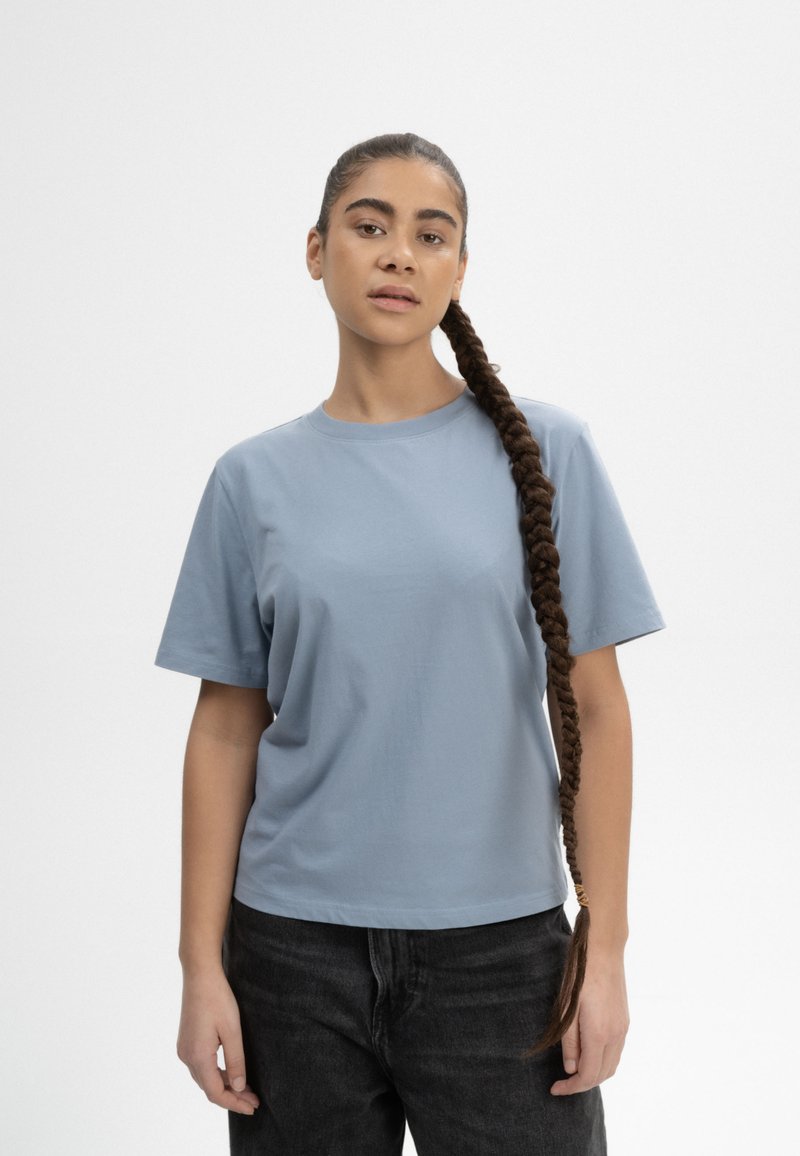 Helles blaues T-Shirt mit kurzen Ärmeln in lockerer Passform, aus weichem Baumwollstoff. Getragen mit dunklen, hoch taillierten Jeans, die eine gerade Silhouette haben.