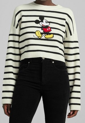 Personne portant un pull court à rayures blanches et noires avec un motif de Mickey Mouse en marche sur le devant, et un pantalon noir.
