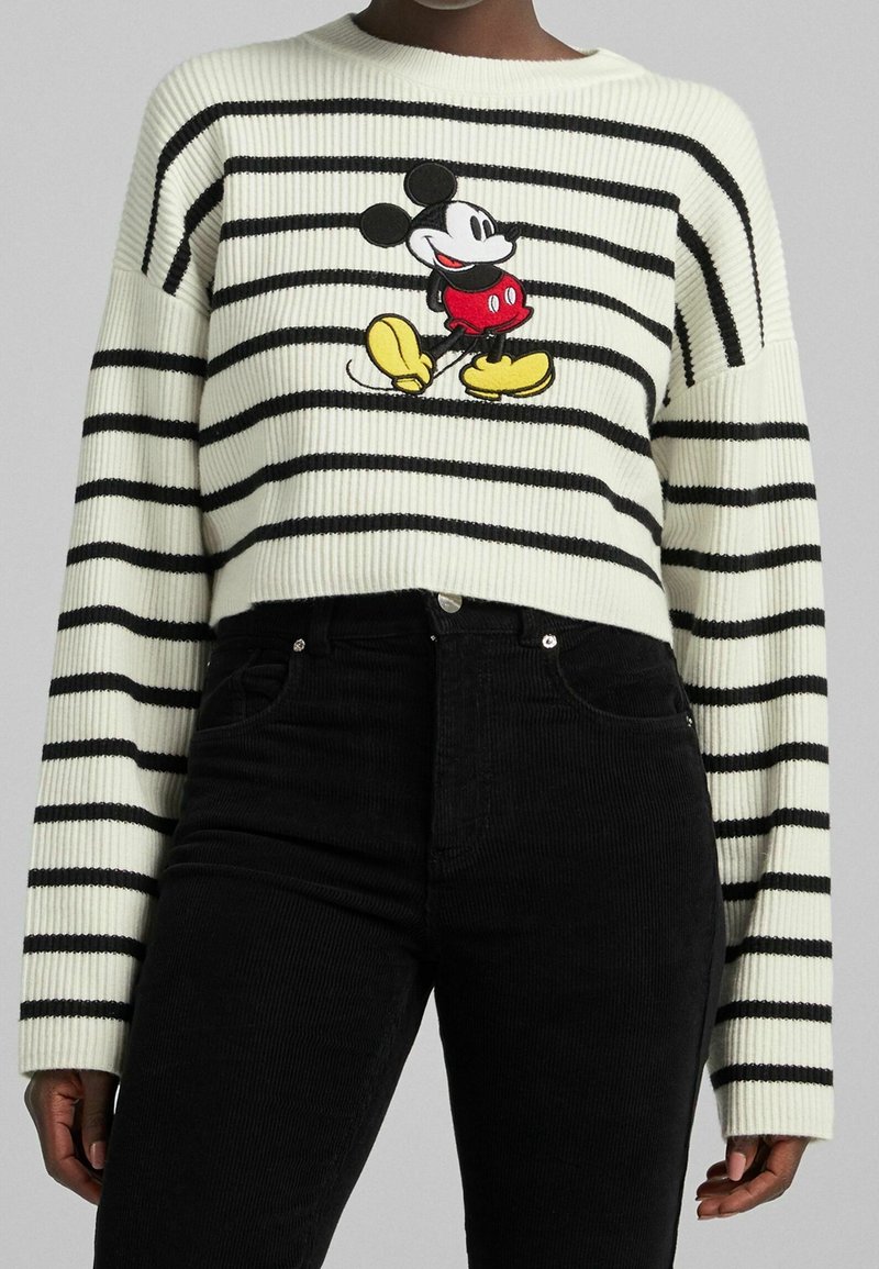 Personne portant un pull court à rayures blanches et noires avec un motif de Mickey Mouse en marche sur le devant, et un pantalon noir.