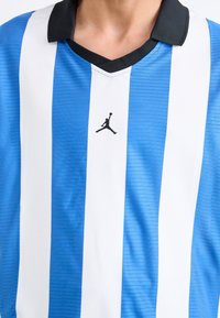 Modrá a biela pruhovaná športová dres s čiernym golierom. Obsahuje malé čierne logo Jumpman v strede. Vyrobený z textúrovanej tkaniny.