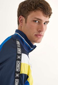 Marinblå zip-jacka med blå, vit och gul accentfärg. Har en hög krage och svart rem med "MUNICH"-logotyp. Slät textur.