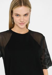 Top nero con maniche trasparenti ricamate che presentano motivi paisley e dettagli di perline. Collo rotondo e vestibilità rilassata.