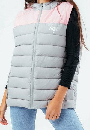 Mouwloos doorgestikt vest met lichtgrijze romp en roze schouderstuk, ritssluiting aan de voorkant, zijzakken, gedragen over een zwart top met lange mouwen en blauwe spijkerbroek.