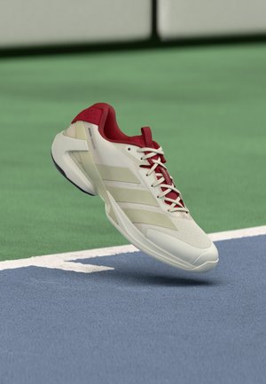 Zapatilla de tenis blanca con acentos rojos, parte superior de malla, tres franjas horizontales, suela plana y cordones visibles, flotando sobre una cancha azul.