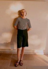 Fiatal nő, rövid szőke hajjal, bézs falnak támaszkodva, fekete-fehér csíkos ingben, fekete rövidnadrágban és barna szandálban.