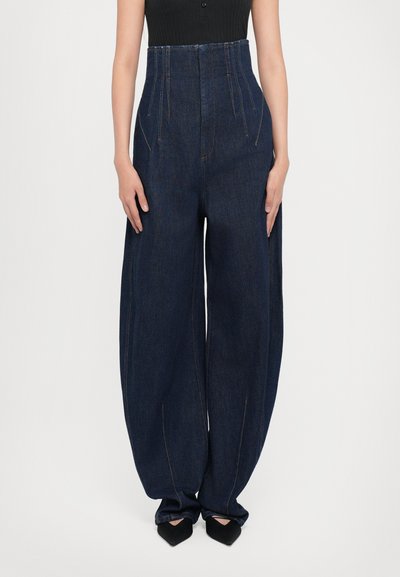 Hoge taille denim broek in donkerblauw, metwijdere pijpen, opvallende naden en een gestructureerde stof. Gecombineerd met zwarte puntige schoenen.