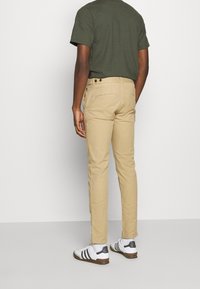 Tan chinos with a slim fit, featuring back pockets and belt loops. Paired with a dark green short-sleeve shirt and white sneakers.