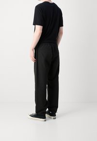 Een persoon die met de rug naar de camera staat, gekleed in een zwart T-shirt met korte mouwen, losse zwarte broek en zwart-witte low-top sneakers, tegen een witte achtergrond.