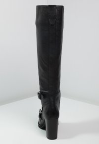 Musta nahkainen polvipituinen saapas, jossa on teksturoitu pinta, paksu korko, säädettävä vyöyksikkö ja tyylikäs siluetti.