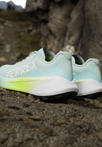 Chaussures de trail bleu clair avec semelles blanches et jaunes reposant sur un sol rocheux près d’une grande formation rocheuse.