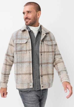 Mand smiler og kigger til venstre iført en beige ternet jakke over en grå skjorte med knapper og en hvid rullekrave, med grå jeans.