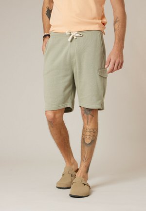 Homme portant un short vert clair à cordon de serrage avec poche latérale et des chaussures slip-on couleur beige, montrant des jambes et des bras tatoués.