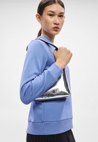 Felpa blu con scollo a girocollo, abbinata a una borsa metallizzata argento lucido tenuta dalla modella, che mette in evidenza linee eleganti e una texture liscia.