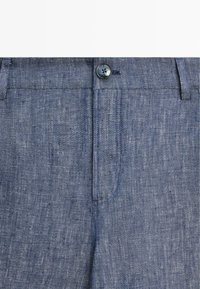 Close-up van blauwe chambray stofbroek met knoopsluiting en zichtbare riemlussen in de taille.