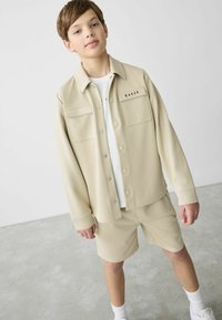 Beige tekstuuri jakke med trykknapper og to frontlommer, båret over en hvit T-skjorte og beige shorts, komplementert av hvite sokker og sko.