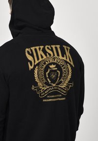 Sudadera negra de algodón, con una gran impresión gráfica dorada en la parte posterior que incluye una corona, una corona de laurel y acentos de texto.