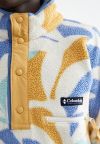 Pull en polaire crème avec des motifs abstraits orange, bleu et vert d'eau. Comprend une poche zippée et une patte de boutonnage jaune. Logo Columbia présent.