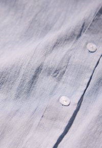 Camicia di lino azzurro chiaro con una texture liscia, pieghe sottili e bottoni bianchi in plastica lungo il risvolto anteriore.