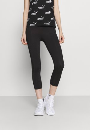 Mustat capri-legginssit, joissa on sileä pinta, yhdistettynä hihattomaan mustaan topsiin, jossa on toistuvia valkoisia logopainoksia. Yhdistettynä valkoisiin lenkkareihin.