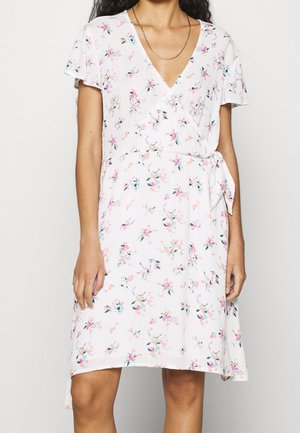 Femme portant une robe portefeuille blanche à fleurs, manches courtes et ceinture nouée sur le côté, longueur mi-cuisse et col en V.