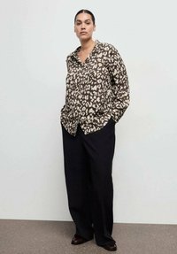 Srajca z gumbi v rjavi barvi z rahlim abstraktnim vzorcem, dolgimi rokavi, sproščenim krojem, kombinirana s temnimi širokimi hlačami, postavljena pred svetlim ozadjem.